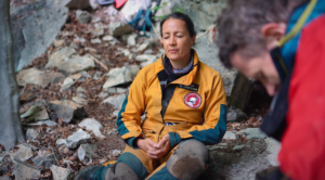 Petra sitzt mit Menschen in der Natur vor einer Höhle und übt die Körperwahrnehmungen.